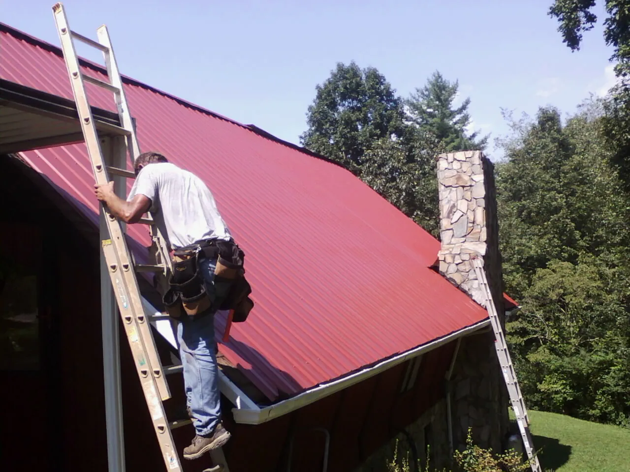 Roofer on a ladder beside a red metal roof and stone chimney — Soffit & Fascia Repair by Roofing Brothers in Mont Alto